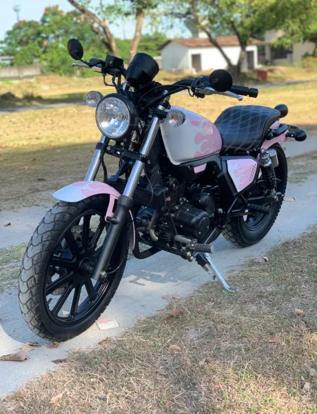 A custom motorcycle with a black and pink design, parked on a grass-lined path. Trees and a small building are in the sunlit background.