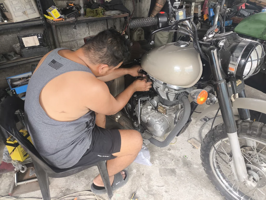 A man in a gray tank top and shorts repairs the engine of a vintage motorcycle in a cluttered garage, conveying focus and concentration.