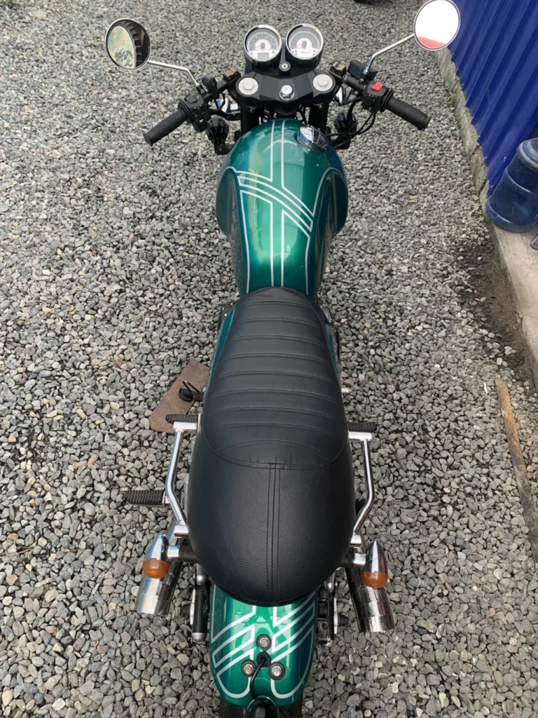 Top view of a green vintage motorcycle with white stripes on a gravel surface. Black leather seat, chrome handlebars, mirrors, and rear lights visible.