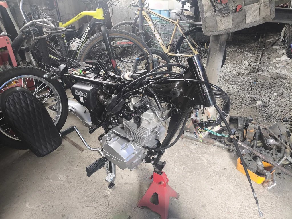 Motorcycle under repair on a concrete floor, frame exposed with engine visible. Bicycles and tools scattered around, conveying a workshop setting.
