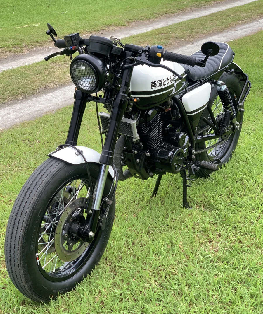 Custom vintage-style motorcycle with white tank, black trim, and retro headlight on grass. Exudes a classic, nostalgic vibe.