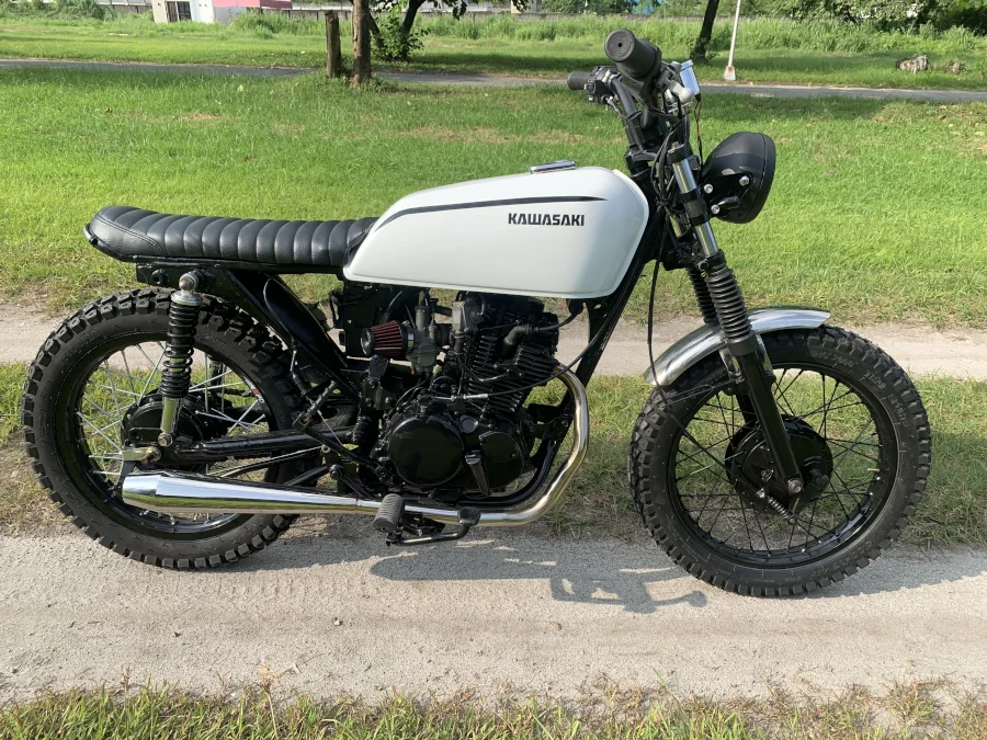 Vintage-style Kawasaki motorcycle with a white fuel tank, black seat, and knobby tires, parked on a path with lush green grass surrounding it.