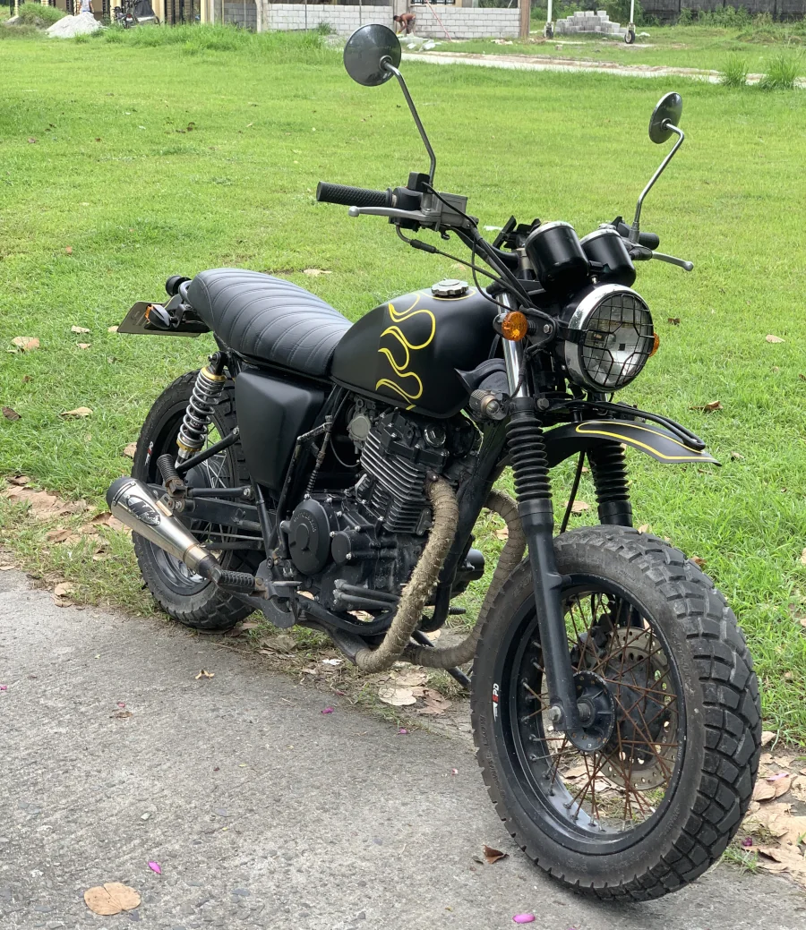 A black motorcycle with yellow flame decals stands on a paved path. It has a vintage design with a round headlight and knobby tires, set against a green lawn backdrop.