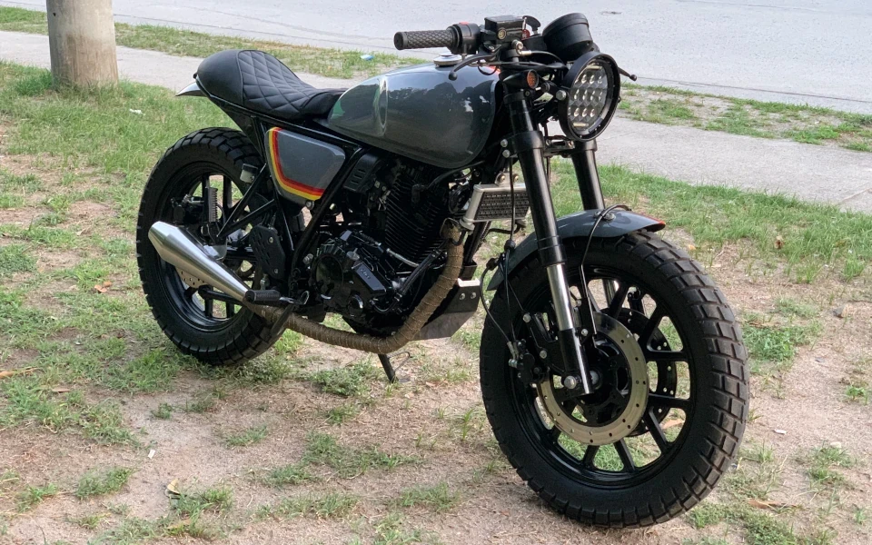 A sleek black cafe racer motorcycle is parked on grass beside a street. It features a retro design and large tires, exuding a vintage vibe.