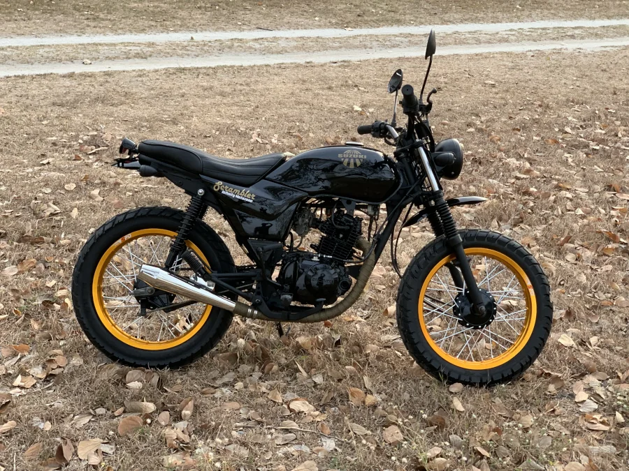 A black scrambler-style motorcycle with yellow rims is parked on dry, leaf-strewn grass. The setting evokes a rustic, autumnal feel.