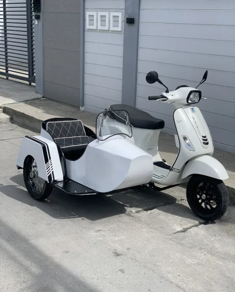 A white scooter with a matching sidecar is parked on a concrete street, next to a modern, gray fence. It conveys a classic, retro vibe.