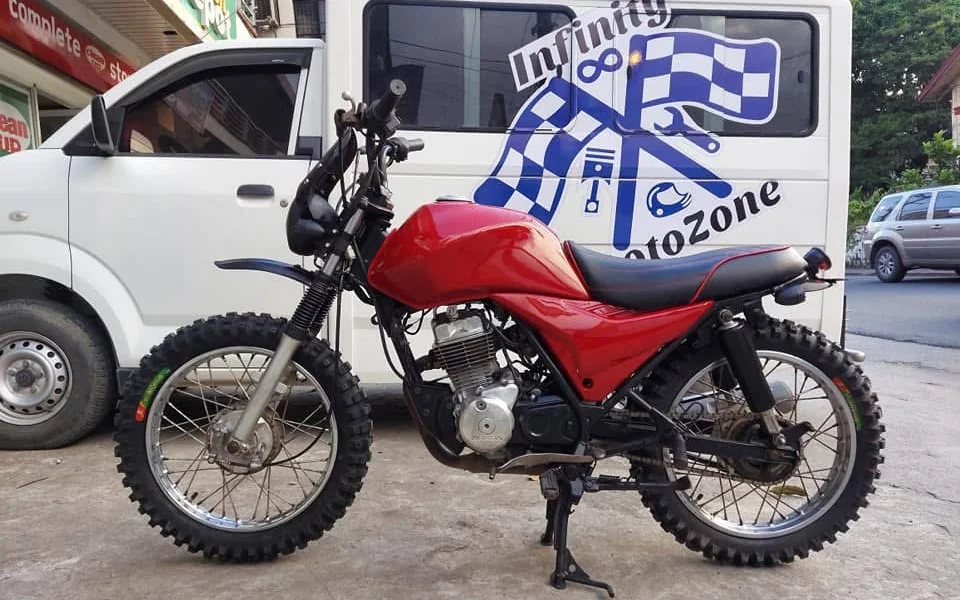 A red dirt bike with knobby tires is parked in front of a white van displaying a blue and white logo with tools and flags. The setting is urban and casual.