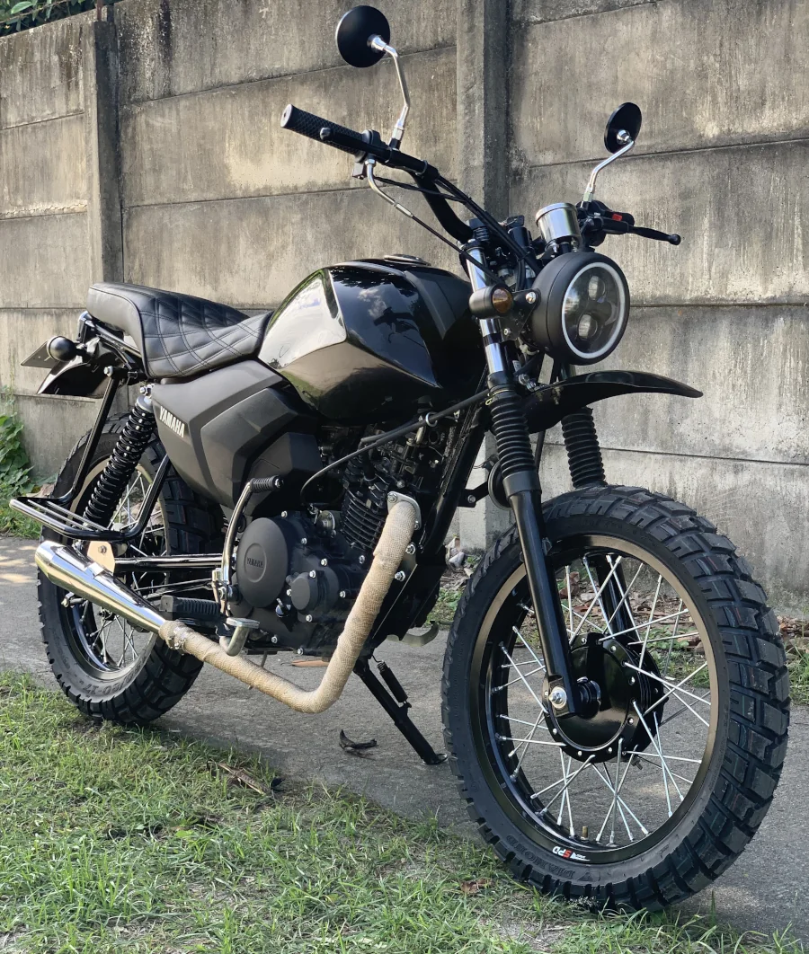 A sleek black motorcycle with knobby tires and a round headlight is parked on grass. The backdrop is a weathered concrete wall, giving a vintage vibe.